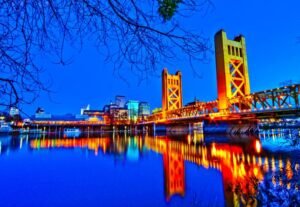Night view of Sacramento’s Tower Bridge lit up in golden lights with city buildings and reflections on the river.